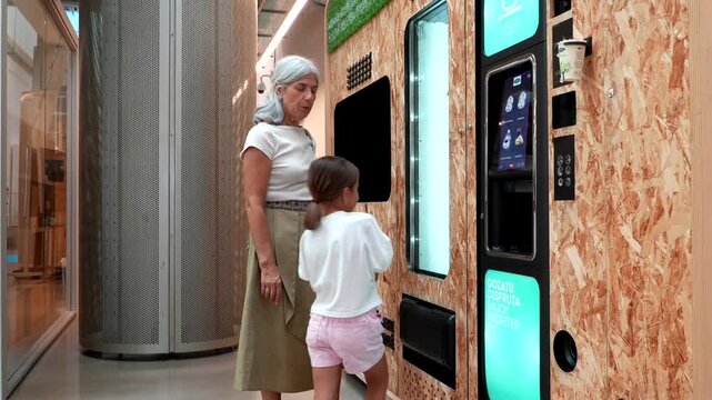 Curious young girl and her elegant grandmother choosing a beverage from a modern vending machine with a touch screen, highlighting intergenerational learning and new technologies