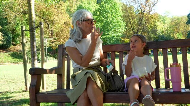 Happy grandmother and granddaughter sitting on a park bench, talking, eating a snack, and enjoying a sunny afternoon together, showcasing a strong family bond and intergenerational connection