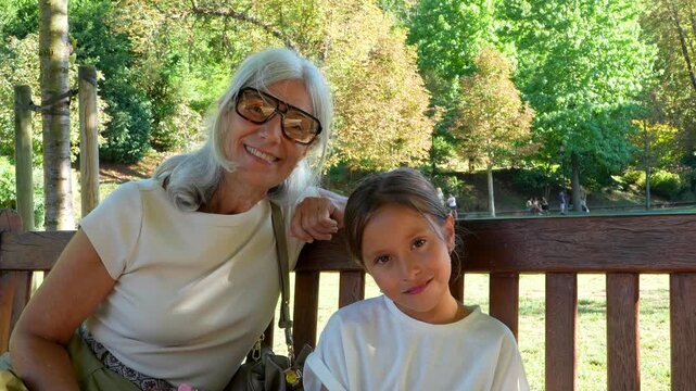 Lovely grandmother and her sweet granddaughter sitting on a wooden bench in a sunny park. Multi generational family members smile and pose for the camera, enjoying their quality time together
