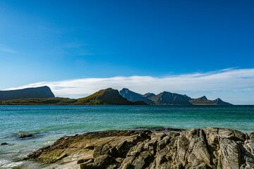 Panoramic view of a rocky coastline with turquoise sea water and majestic mountain peaks under a clear blue sky in Norway with copy space © Quality by Simon