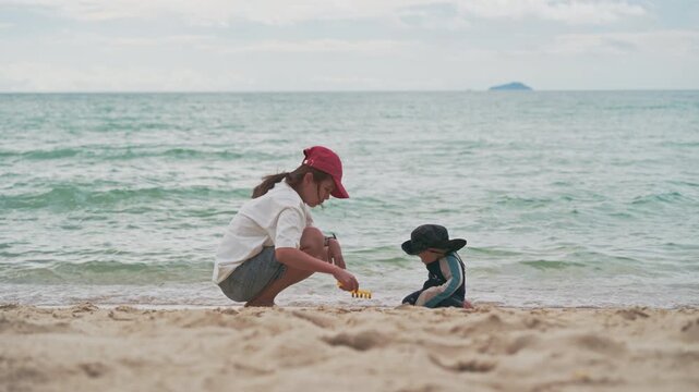 Happy mother and adorable baby boy sitting on sandy beach together during summer vacation in Thailand.