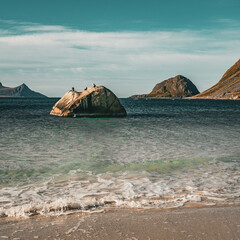 Vertical view of a scenic ocean beach with a solitary rock in the sea and mountain peaks under a calm blue sky in Norway with copy space © Quality by Simon