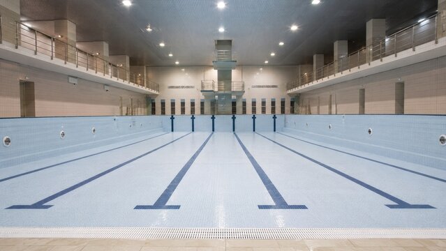 Drained indoor Olympic-size swimming pool with tiled lanes, lane markings, diving platform, spectator balconies, and overhead lighting