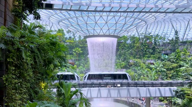 Jewel Changi airport indoor waterfall and skytrain passing through lush green tropical garden in Singapore.