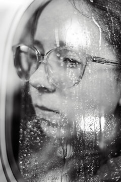 Pensive woman with glasses behind rainy window