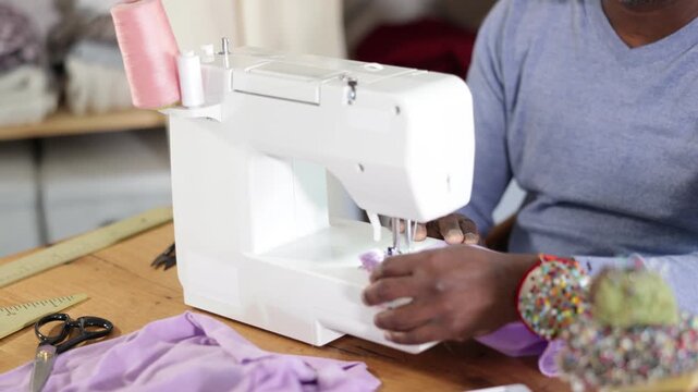 Close-up of hands of professional black male tailor working at sewing machine, stitching beige linen (soft lilac) fabric with precision at table in workshop 