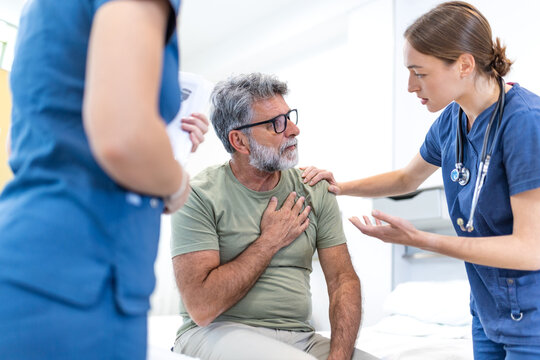 Heart pain and medical examination. Senior man sitting on hospital bed holding chest while doctor examines him, Healthcare and medicine concept, Patient consulting physician about cardiac problem.