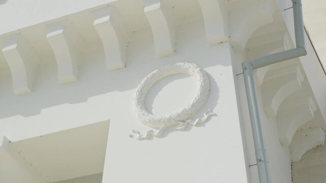 Oval decorative relief sits beneath white cornice near drainpipe. Sculpted frame projects from wall while highlights restored facade detail