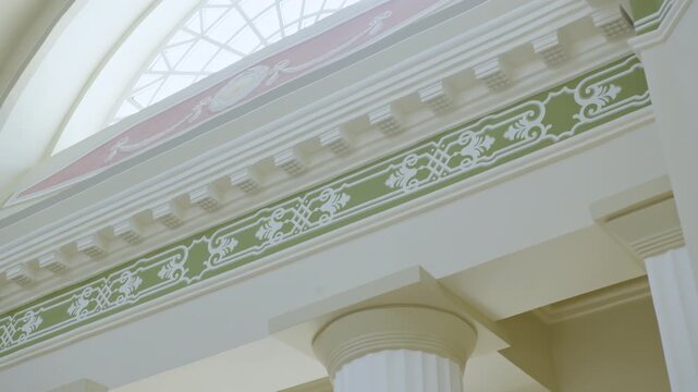 Decorative frieze runs above white columns beneath glass roof. Ornamental border adds detail while highlighting classical interior design