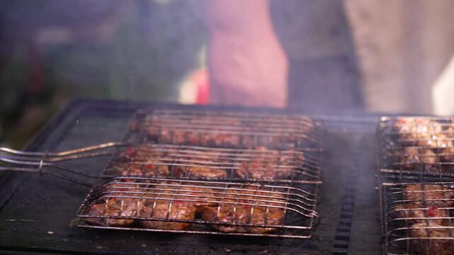 Close up of a chef flipping a wire grilling basket full of meat patties on a smoky grill