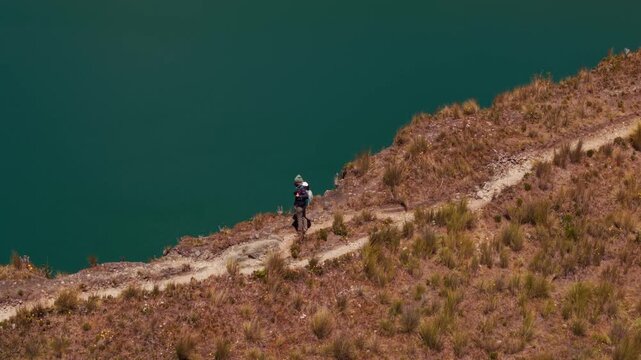 Traveler with a backpack hiking on a narrow path along the volcanic crater lake of quilotoa, ecuador