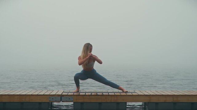 Athletic flexible Caucasian woman exercising and stretching doing Cossack squats on a wooden deck beside a misty lake, showcasing strength, mobility, balance and functional fitness.