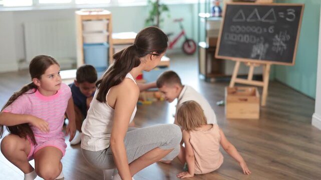Group of children of different ages listening to teacher and repeating movements, walking like ducklings in modern childcare center 