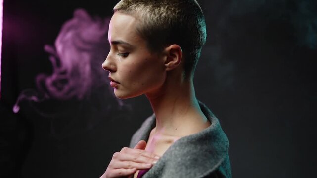 Camera starting, sliding buzzcut woman tracing neck, chest in studio portrait with gray coat, smoke