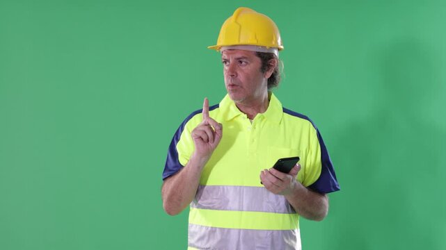 Adult male worker wearing reflective polo shirt and safety hard hat stands against green screen, holding smartphone and checking inventory 