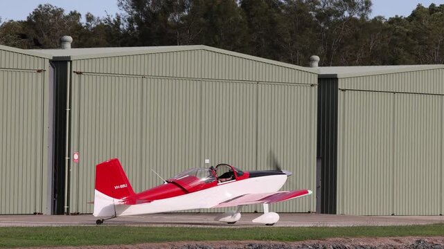 Small red and white airplane on tarmac in front of hangar with propeller spinning.