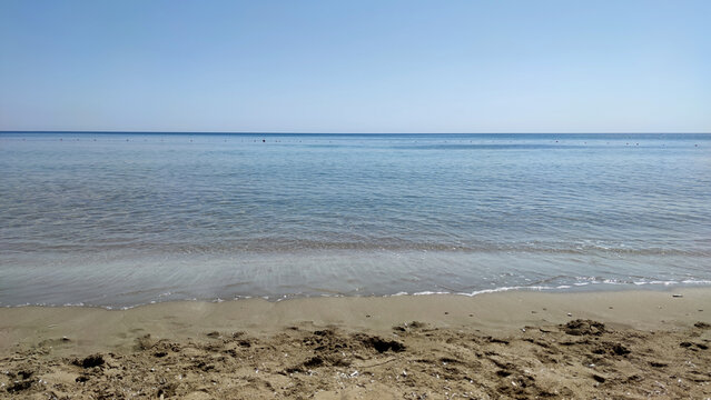 A wide eye-level view of a calm sandy beach with gentle waves meeting the clear blue sea under a cloudless sky in Cyprus.
