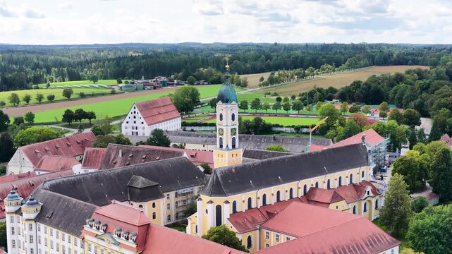Malerische Luftaufnahme von Ochsenhausen mit der barocken Klosteranlage, eingebettet in die idyllische, gr&uuml;ne H&uuml;gellandschaft Oberschwabens unter blauem Sommerhimmel mit Wolken.