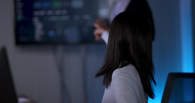 Adult female technology professional works at computer workstation, turns toward presenter near digital dashboard and listens while reviewing software data during collaborative strategy meeting.