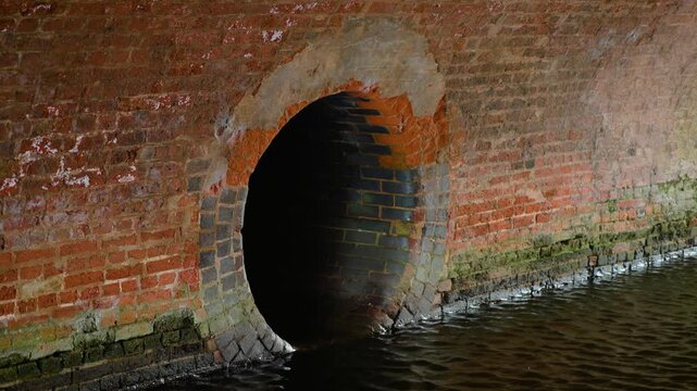 Wastewater drainage outlet in old brick wall beside waterway. Sewer discharge opening beneath weathered masonry with moss at waterline. Environmental pollution from urban drainage infrastructure near