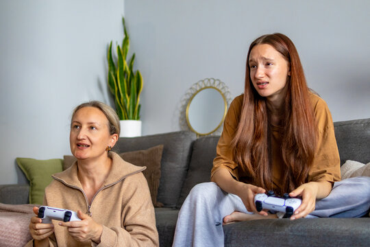 Cheerful mother talking while her disappointed teenage daughter loses a competitive video game. Concept of playful rivalry, gaming competition, and different generational reactions during family play.