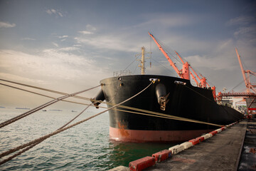 Fototapeta premium Cargo ship moored at industrial harbor with orange crane, heavy rope, calm seawater, and dramatic cloudy sky creating maritime transport scene