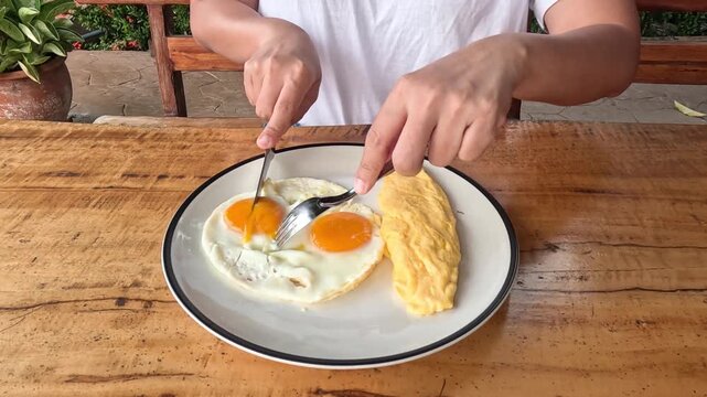 Woman hands cutting runny fried egg yolks with knife and fork during breakfast at wooden table.