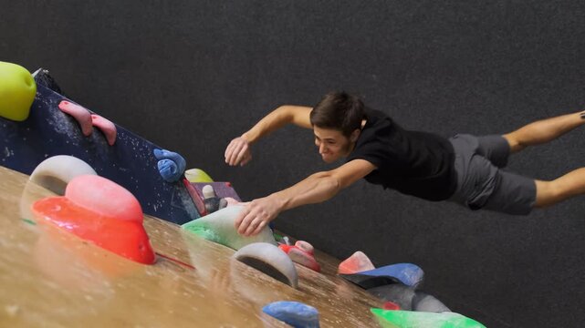 Beginner guy attempts to scale wall by gripping hooks slow motion. Participant falls on floor unable to conquer challenging wall