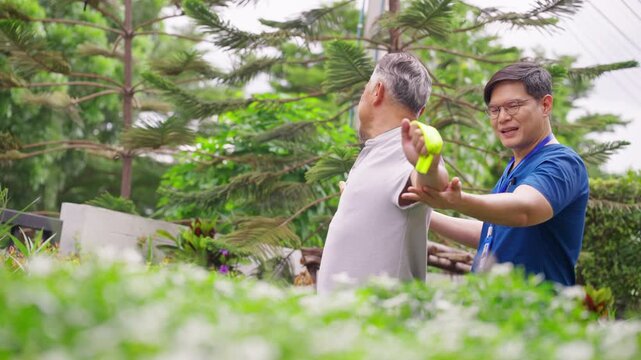 Asian male caregiver or nurse helping senior man exercise with resistance band in garden at home, supporting strength, mobility, rehabilitation, healthy aging, physical fitness and elderly wellbeing