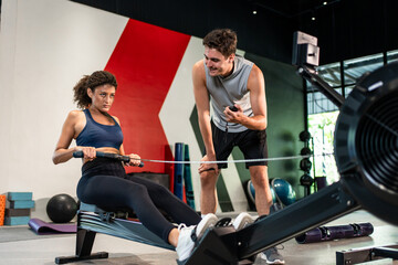 Fototapeta premium Young athletic woman using rowing machine with trainer in fitness gym. 