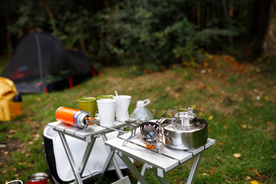 High angle shot of a camping picnic setup with stainless steel kettle, gas stove, canisters, and cups on two aluminum folding tables, with a tent and forest in the blurred background