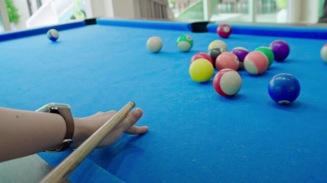 Close up of hand aiming cue on blue pool table near racked balls. Focused player lining up a break shot in a bright sunlit game room with colorful billiard balls on the table near windows.