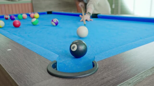Close up of black eight ball on blue pool table near pocket. Attractive player in the background lines up a precise cue shot, creating a relaxed recreational vibe in a bright game room at home.