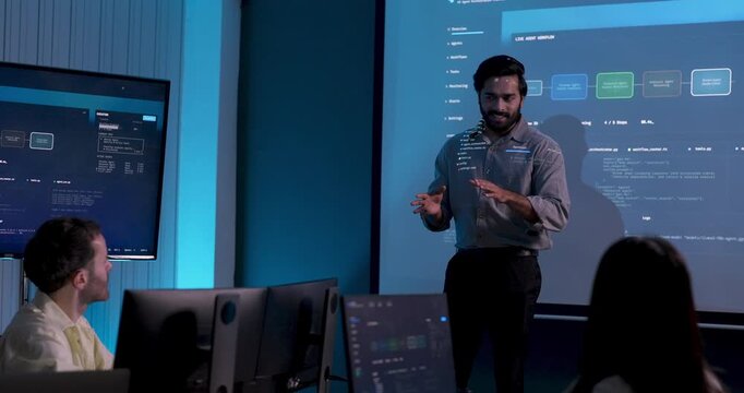 Adult male technology professional speaks to seated business team, gestures beside software dashboard and explains system data, digital workflow and strategy during collaborative office meeting.