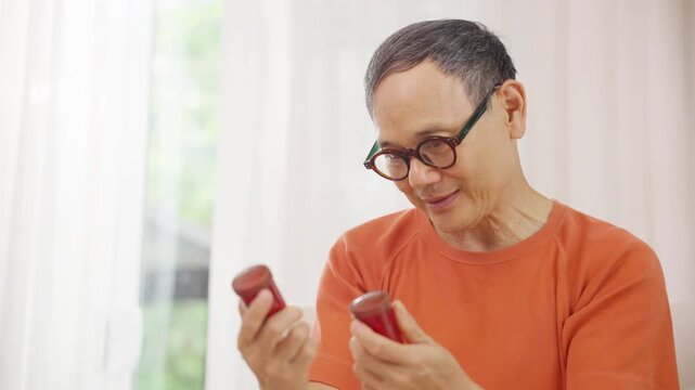 Asian senior man taking medicine from pill bottle at home, elderly healthcare with medication management, daily treatment and prescription drugs, health support and chronic disease care.