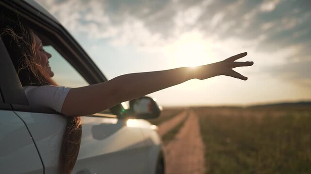 Car rides road with girl leaning out window. Hand waves into sky at sunset. Woman enjoys freedom of motion. Road leads through golden light. Travel moment for girl. Hand flows with freedom wind.