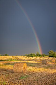 Countryside field with hay bales under a warm sunset rainbow.