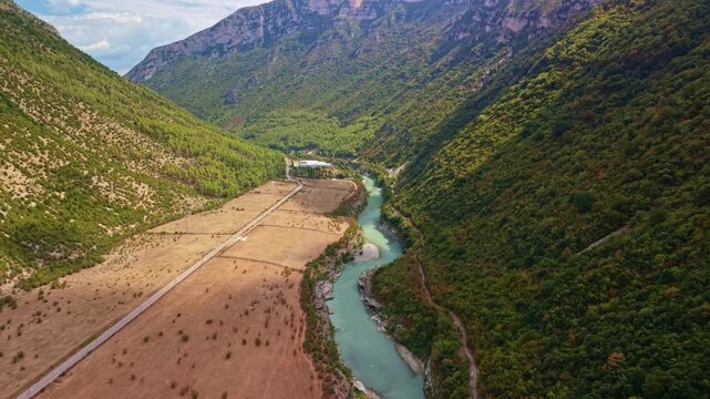 Vjosa River meanders below rugged peaks, cutting narrow path through forested slopes and open plain. Elevated view presents peaceful river corridor with dramatic alpine surroundings.
