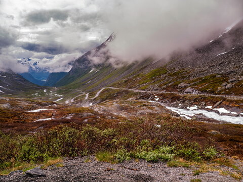 Stryn, Norway - 11.06.2014: Winding road through a dramatic mountain valley under heavy low clouds with a rushing river and snow patches