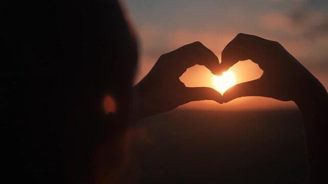Heart shape in hands glows in sunset. Silhouette hands express emotion of love. Love and light form symbol together. Sunset gives shape meaning. Emotion in silhouette hands holding love and light