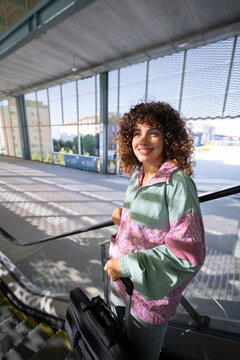 White woman holding luggage on escalator in urban outdoor setting