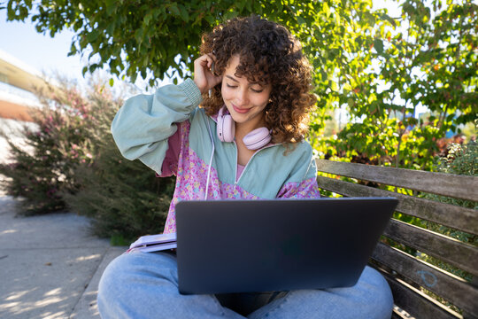 White woman sitting on a bench outdoors using laptop with headphones