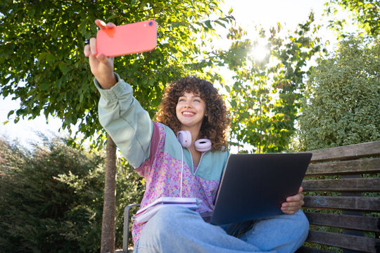 White woman taking selfie outdoors on bench with laptop and notebooks