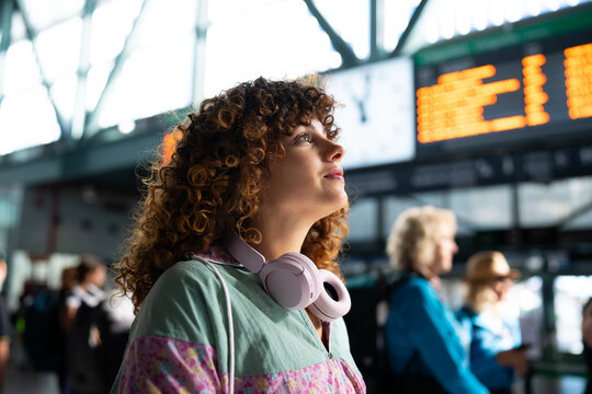 White woman with headphones looking at train arrival board in station