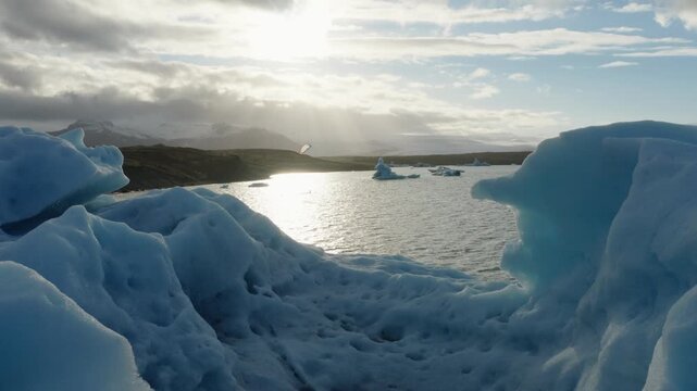 Large blue iceberg floats in Jokulsarlon Glacier Lagoon with smaller ice formations and snow covered mountains beyond, southeastern Iceland, drone push in and pass over.