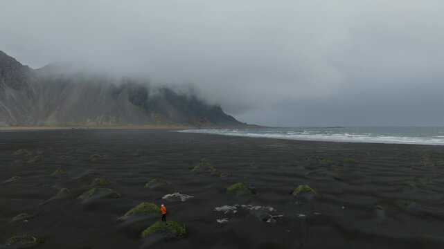 Black volcanic beach extends beneath fog covered Reynisfjall mountain with moss covered sand hummocks and Atlantic surf, Iceland, drone pullback.