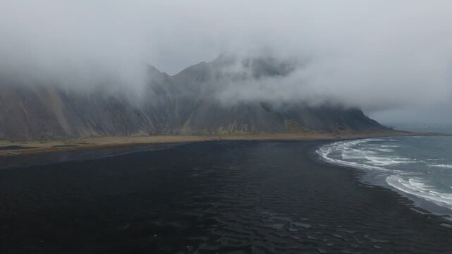 Reynisfjara black volcanic beach curves beneath Reynisfjall tuff ridge, with Atlantic surf and low orographic cloud obscuring the peak, Iceland, drone forward flyover.