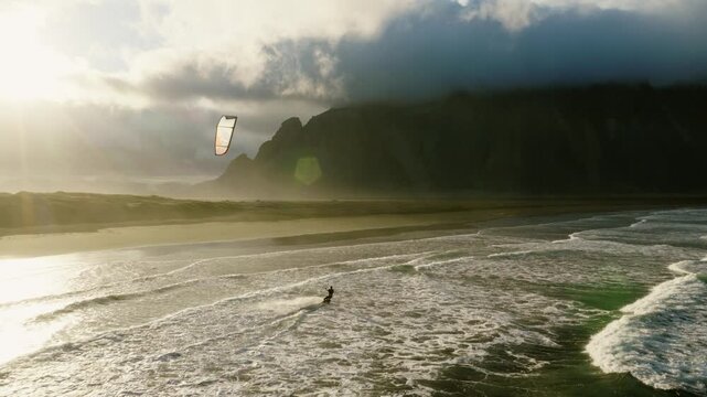 Kitesurfer tracks across Atlantic surf beside black volcanic sand beneath steep Reynisfjall mountain and low cloud, South Coast Iceland, drone tracking.