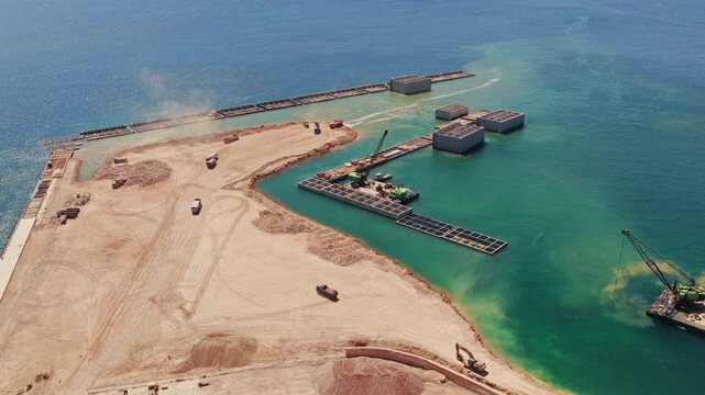 Trucks and excavators moving earth on a massive coastal engineering project, creating a new harbor with concrete caissons and breakwaters extending into the turquoise sea under a clear blue sky
