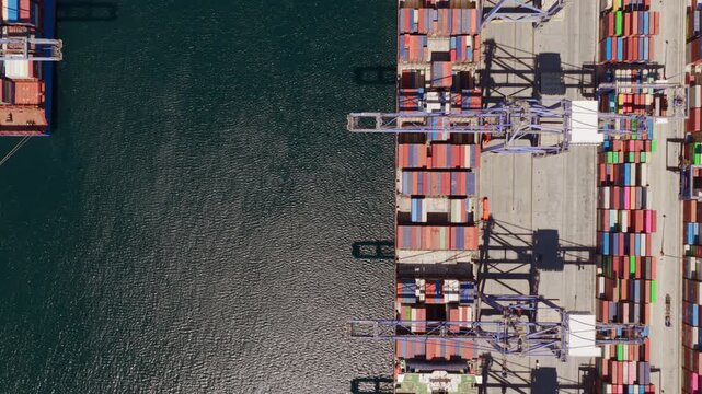 Top-down aerial view showing a large cargo container ship departing from a commercial port terminal, revealing the industrial harbor and rows of colorful intermodal containers on the quay